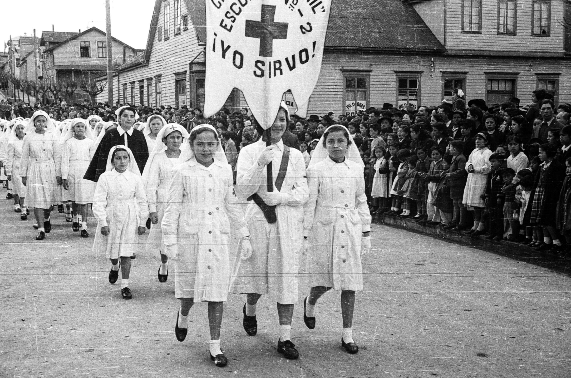 plano general de un grupo de Niñas desfilando vestidas de blanco un velo en sus cabezas, en el frente una de las niñas carga un pendón que tiene escrito Cruz Roja Juvenil, Escuela Niño, ¡Yo sirvo!'. En un costado de la calle, sobre la vereda, hay un grupo de Niños y Niñas junto con Hombres y y Mujeres observándolas mientras desfilan por la calle