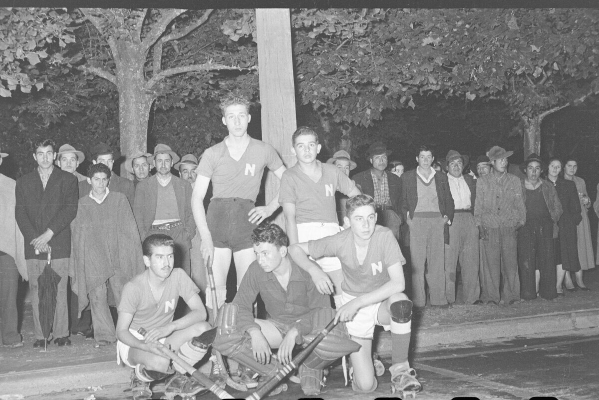 Retrato grupal de cinco jugadores de hockey posando frente a un poste, en el fondo hay una hilera de hombres y mujeres observando de pie.