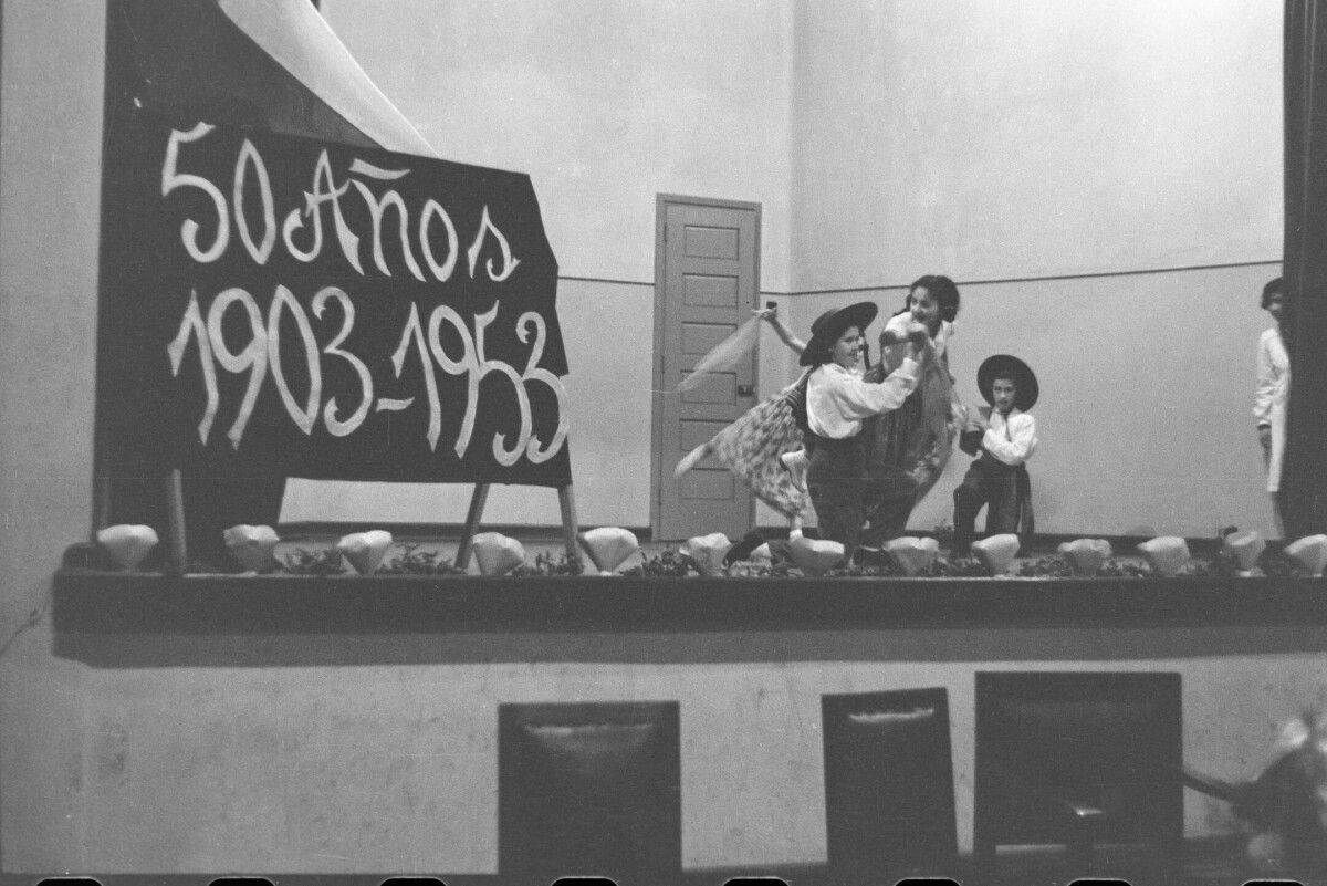-plano general de dos niñas y dos niños bailando cueca sobre un escenario, caracterizados de acuerdo a la vestimenta típica chilena de la --China y el huaso-- chileno. En un costado del escenario hay cartel puesto, el cual dice --50 años. 1903 - 1953-- y en el otro costado hay una persona parada, observando detrás del telón. Los niños y niñas están realizando la presentación para el aniversario de la escuela superior de niñas Nº2.-
