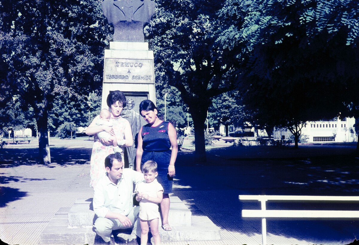 Retrato de plano entero de dos mujeres con Vestido una de ellas con un Bebé en brazos, un hombre con Traje y un niño, todos posando de frente junto a estatua de Teodoro Schmidt en Plaza de Armas de Temuco