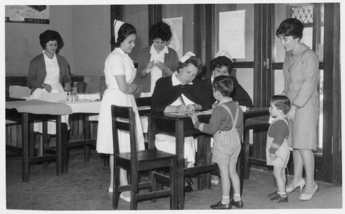 plano general de una enfermera, sentada junto a una mesa, anotando en un libro mientras un niño se encuentra parado frente a ella. A su alrededor se encuentra una mujer junto a otro niño y otras cuatro enfermeras, una de ellas es Mirna Perez Diaz.