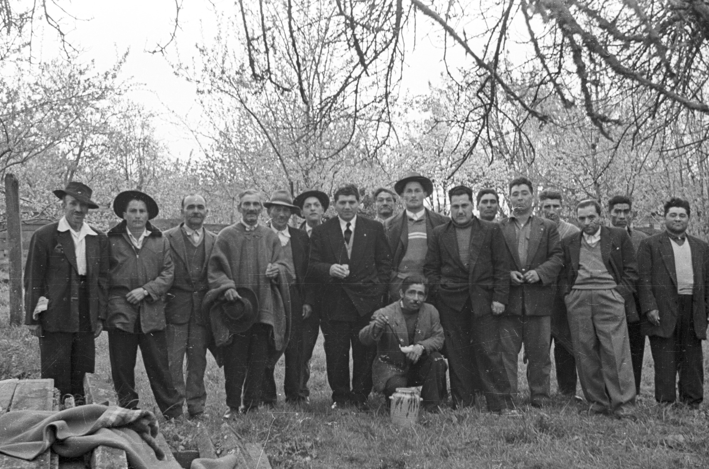 Retrato grupal de plano entero de Hombres de Traje, sombrero o manta, posando formados en una hilera. Hincado, junto a ellos hay un hombre posando con un cucharón en la mano. Se encuentran al aire libre en un lugar no identificado