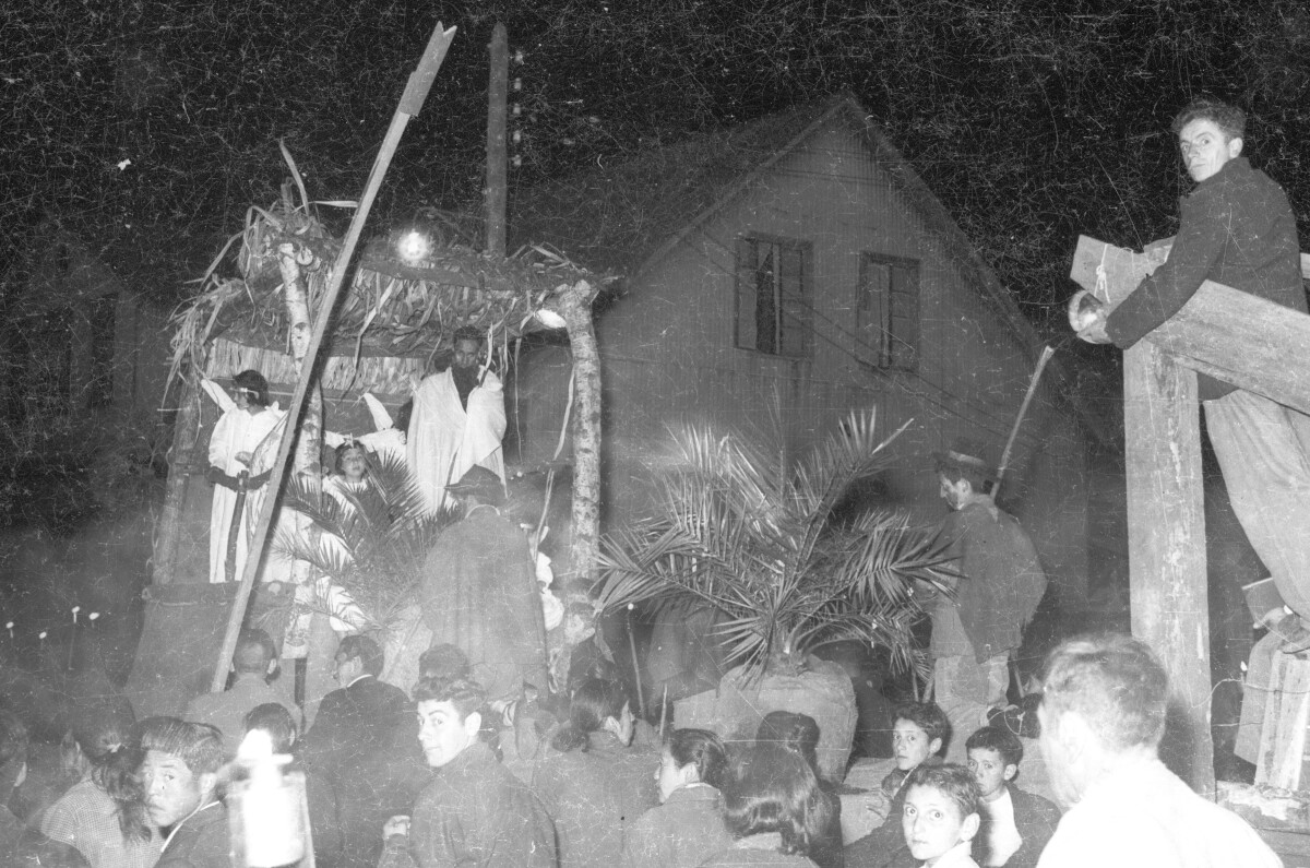 plano general de un grupo numeroso de personas, siguiendo a un carro alegórico donde hay un grupo de niñas disfrazadas de ángeles. el carro está decorado como un pesebre. Se encuentran desfilando durante la noche por las calles de La Unión
