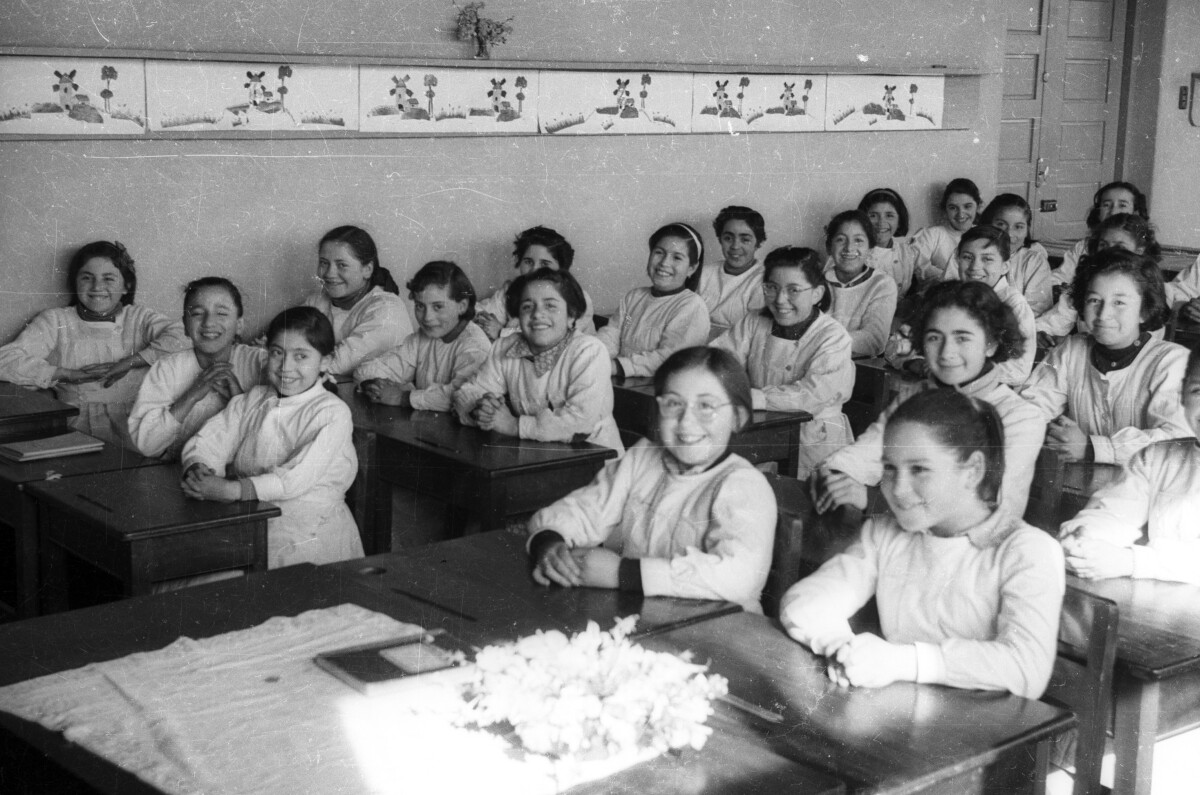 Retrato grupal de plano entero de grupo de niñas con delantal sentadas en sus pupitres en una sala de clases