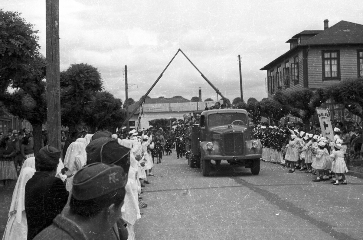 plano general de un grupo de niñas, Mujeres y Hombres de la cruz roja, lanzando serpentina a un camión, en el cual viene Carlos Vogel parado en la parte trasera, durante un homenaje realizado por su llegada a la ciudad de La Unión