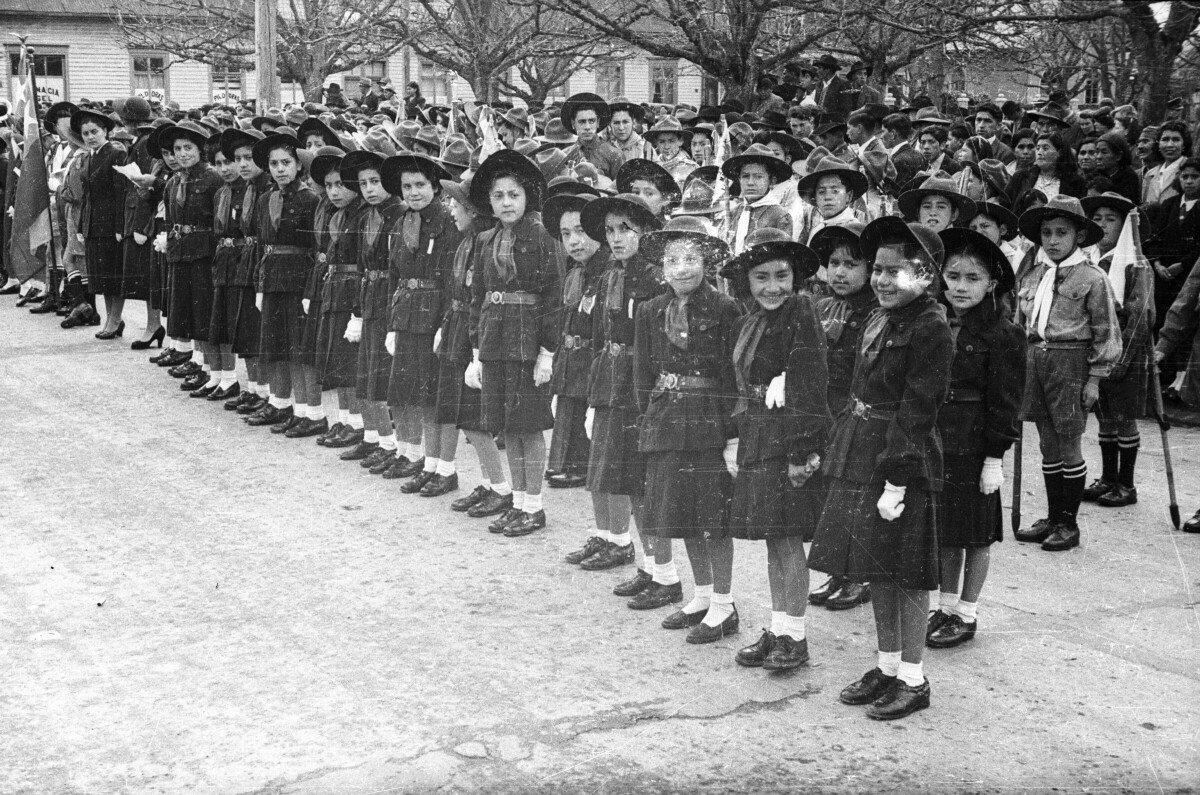 Plano entero de un grupo de niñas y niños de la brigada de scout formados en hileras. En el frente hay dos hileras conformadas por la brigadas de scout femenino, todas ellas llevan puesto su uniforme al igual que los Niños ubicados en dos hileras tras ellas, algunos de ellos llevan banderines y dos llevan la bandera chilena en el final de la primera fila. Junto a ellos, sobre la vereda, se encuentras Mujeres, Hombres y Niños observándolos desde La Plaza de La Concordia de La Unión