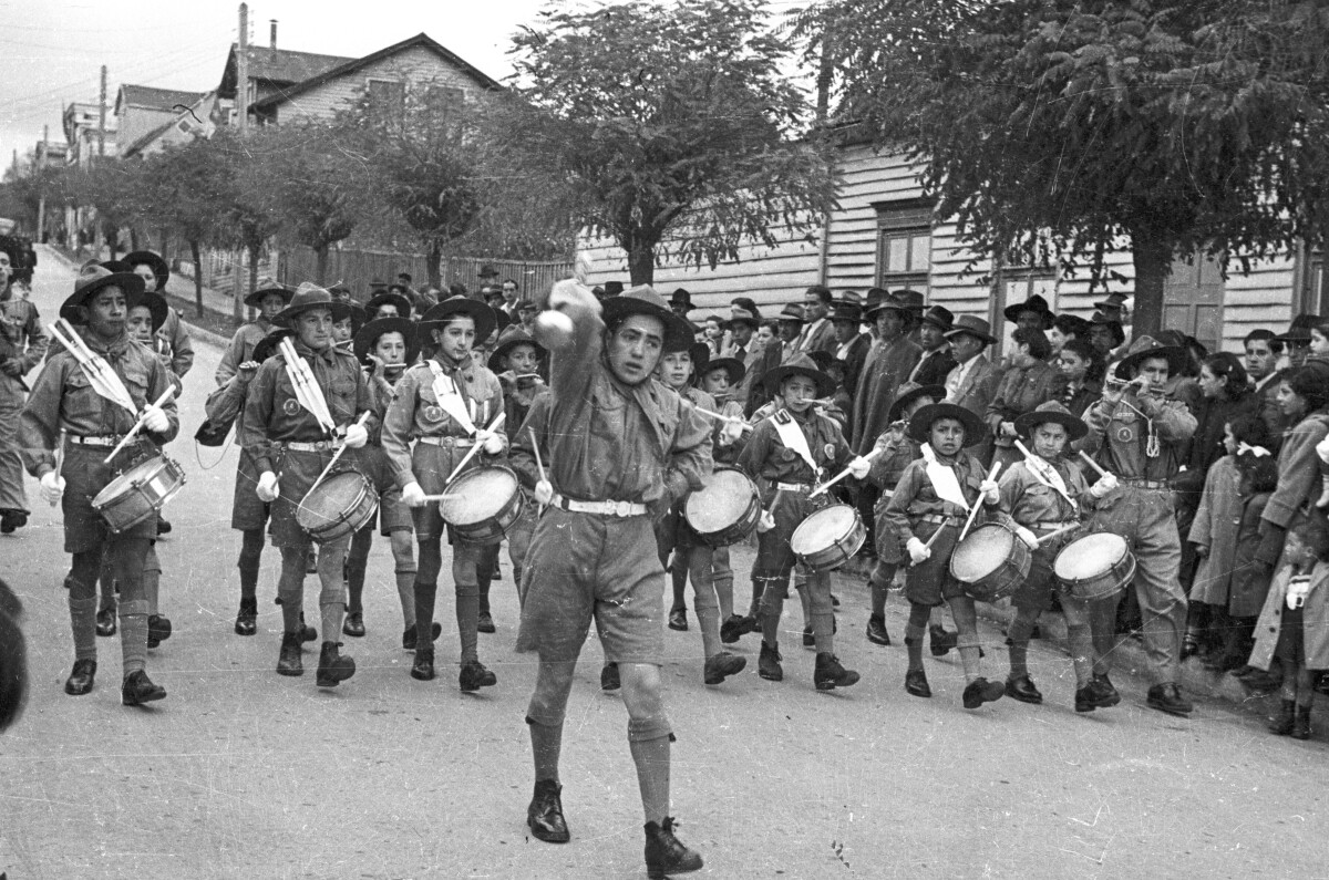 Plano entero del tambor mayor moviendo su guaripola mientras la orquesta sinfónica infantil le sigue tocando sus instrumentos. Todos pertenecen a una brigada de scout no identificada y a un costado, un grupo de Mujer, Niñas y Hombres los observa desfilar por lo largo de una calle pavimentada