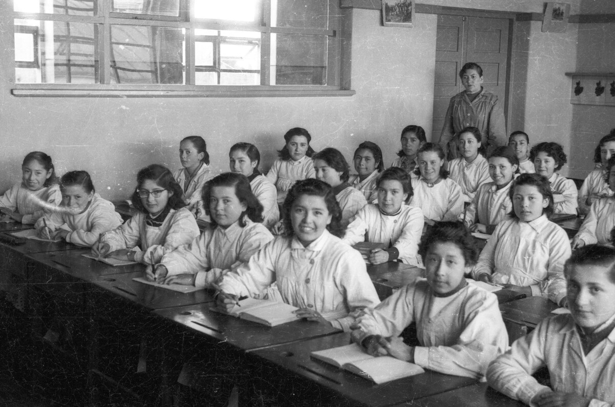 plano general de un grupo de niñas escribiendo en sus cuadernos, sentadas junto a sus pupitres. En el fondo, se encuentra la profesora parada junto a la puerta de la sala de la escuela superior de niñas Nº3