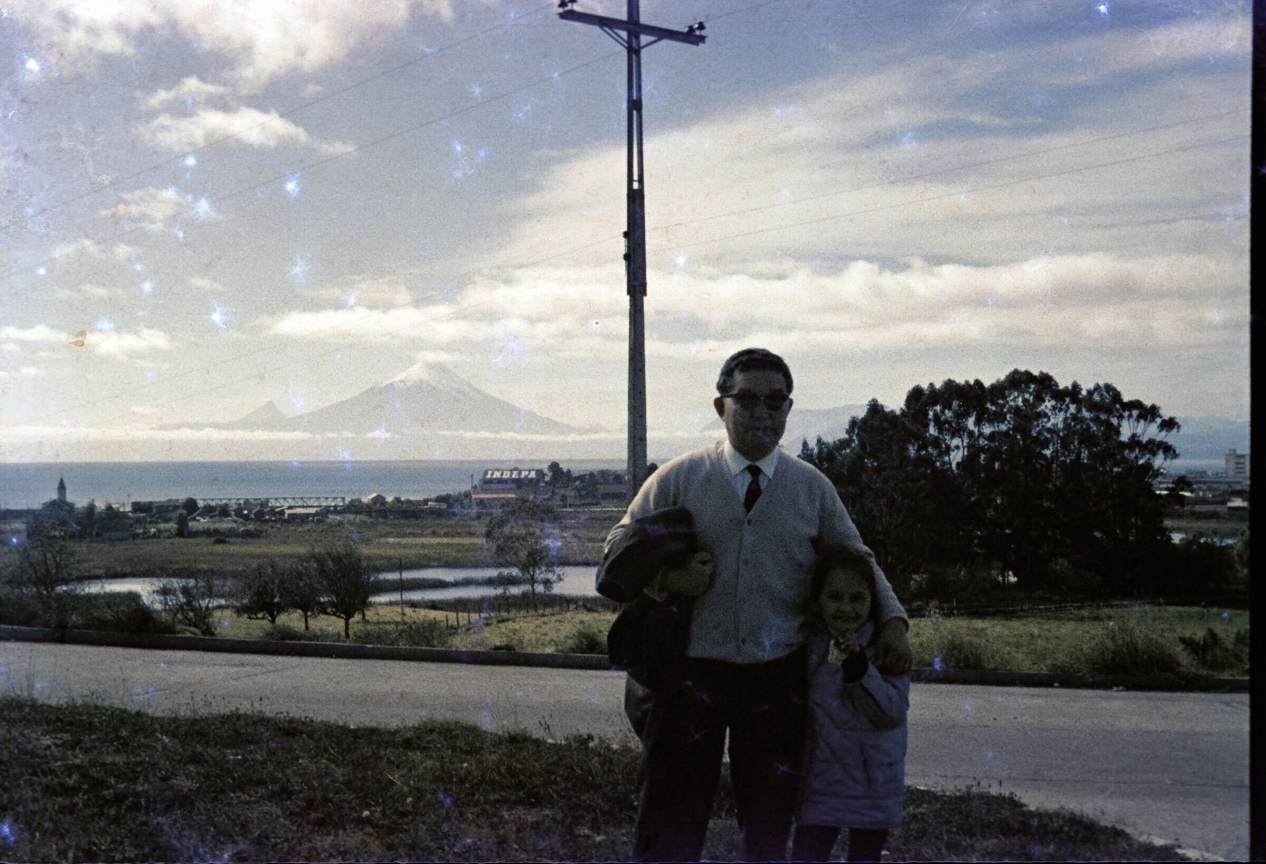 Retrato plano americano de Hombre con Traje abrazando a niña. Detrás de ellos un paisaje con árboles, el lago Llanquihue y los volcanes Osorno y Puntiagudo.