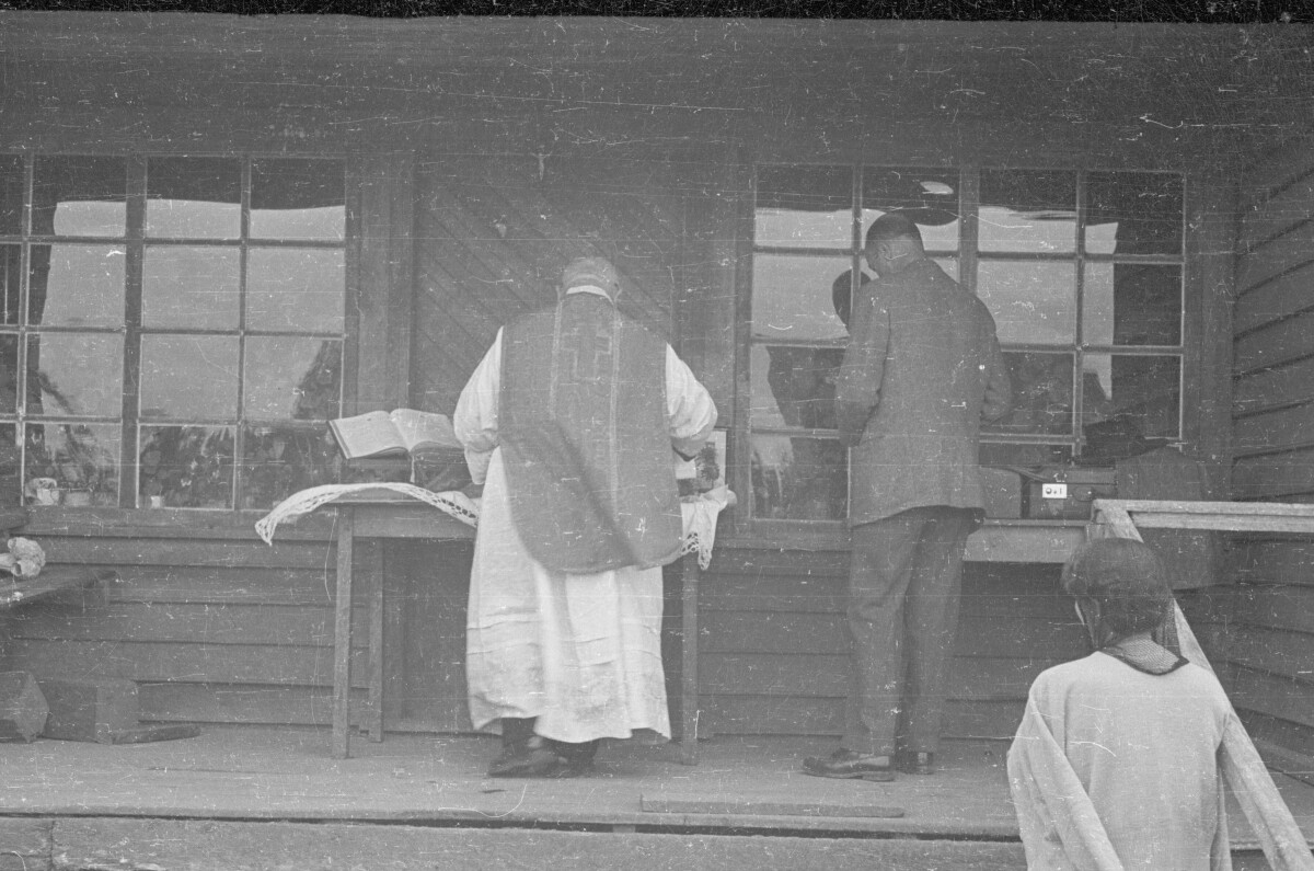 Plano general de sacerdote de espaldas preparando el sacramento, a su lado lo acompaña un hombre de traje también de espaldas.