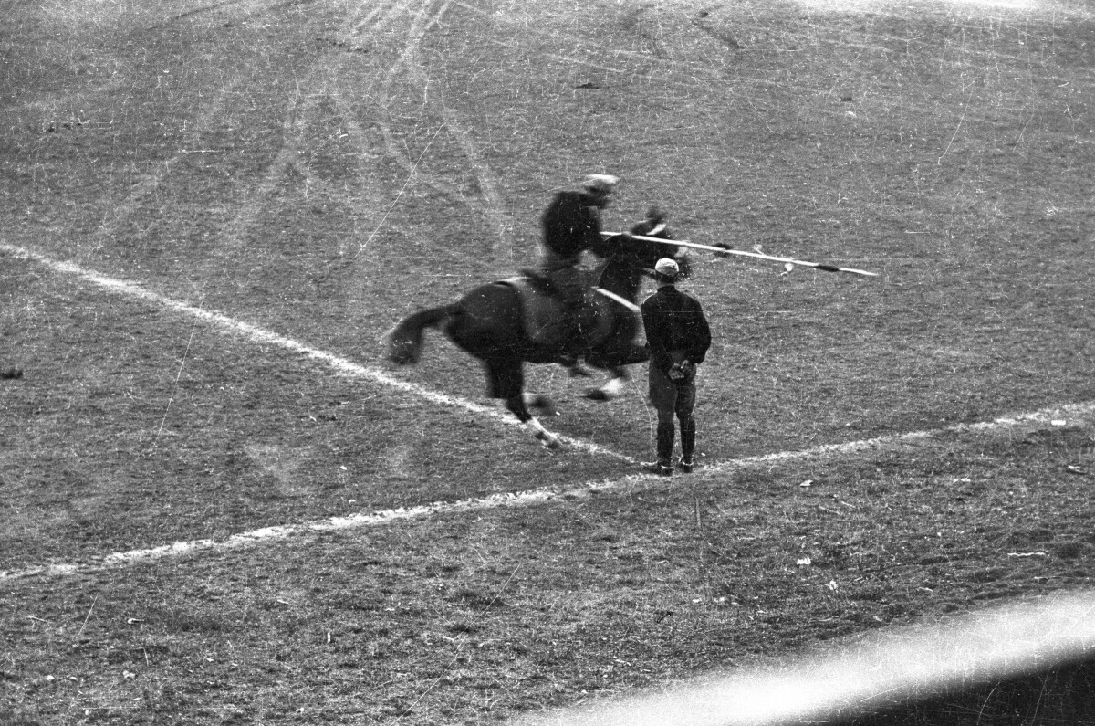 Plano entero de un hombre parado junto a un hombre galopando sobre un caballo mientras realizan una presentación en el interior de la cancha ubicada en el estadio Carlos Vogel