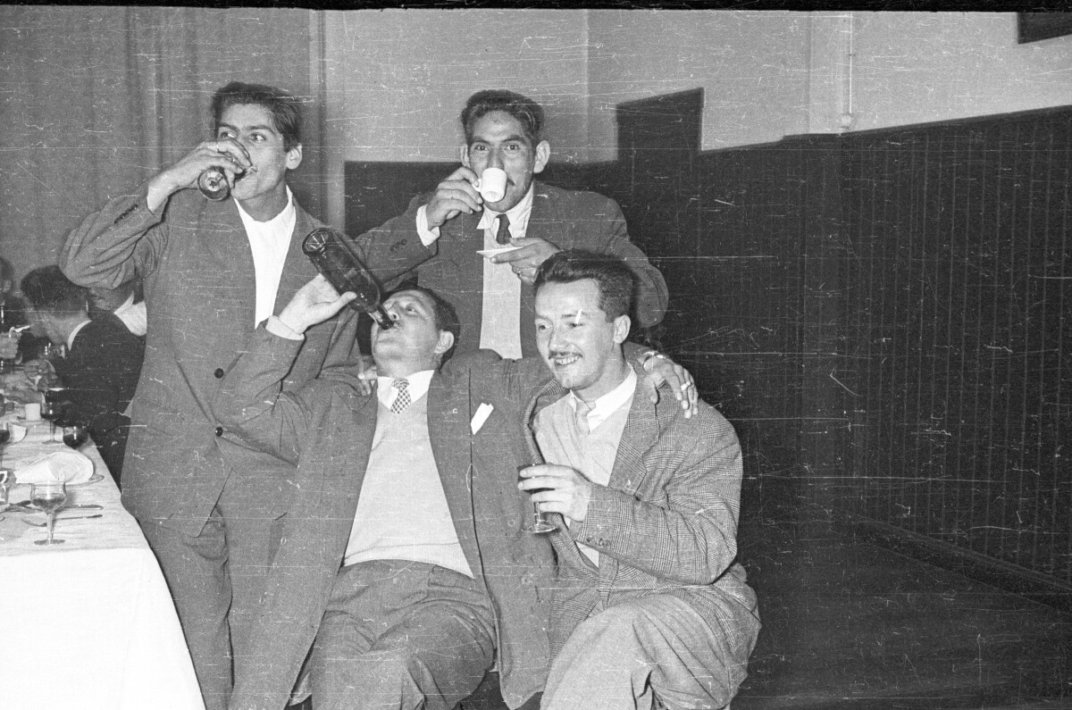 Retrato de plano entero de cuatro hombres posando, dos están de pie y dos sentados, dos de ellos está tomando una copa, otro una taza y otro desde una botella, están junto a una mesa durante una celebración en el interior de un local