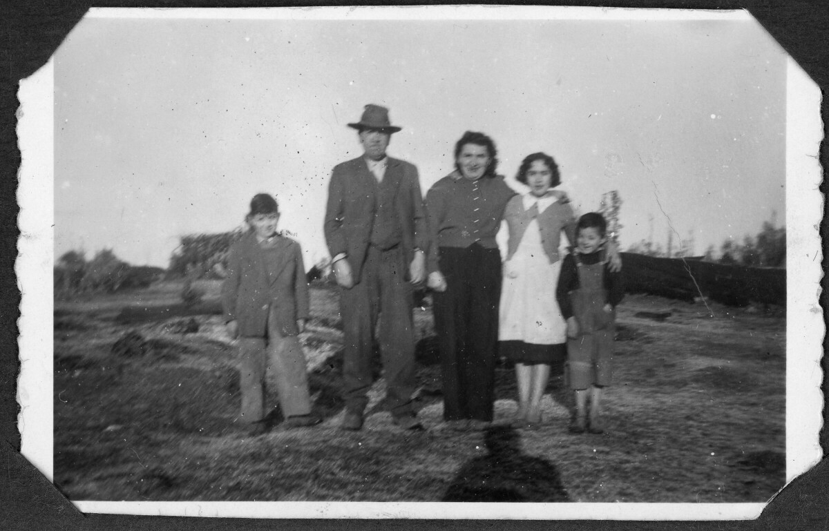 Retrato grupal de cinco personas. Un hombre adulto, una mujer adulta, una mujer joven y dos niños. Están en un ambiente rural. La mujer viste pantalón y blusa, la mujer joven un vestido con chaleco, el hombre adulto viste terno y sombrero, uno de los niños viste terno, y el otro una jardinera.
