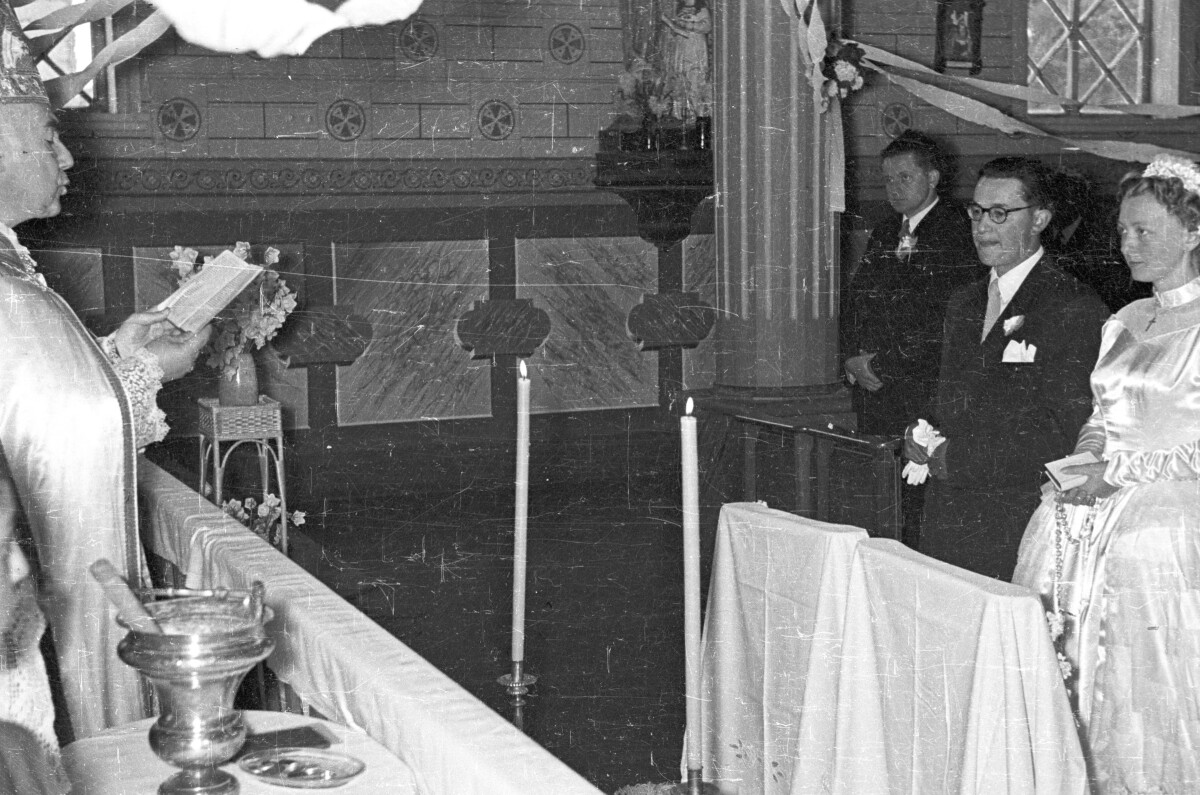 Plano entero de un sacerdote leyendo la biblia frente a una pareja de novios, quienes se encuentran parados por detrás de unos reclinatorios. Están en el interior de una parroquia no identificada, durante una misa de celebración de matrimonio