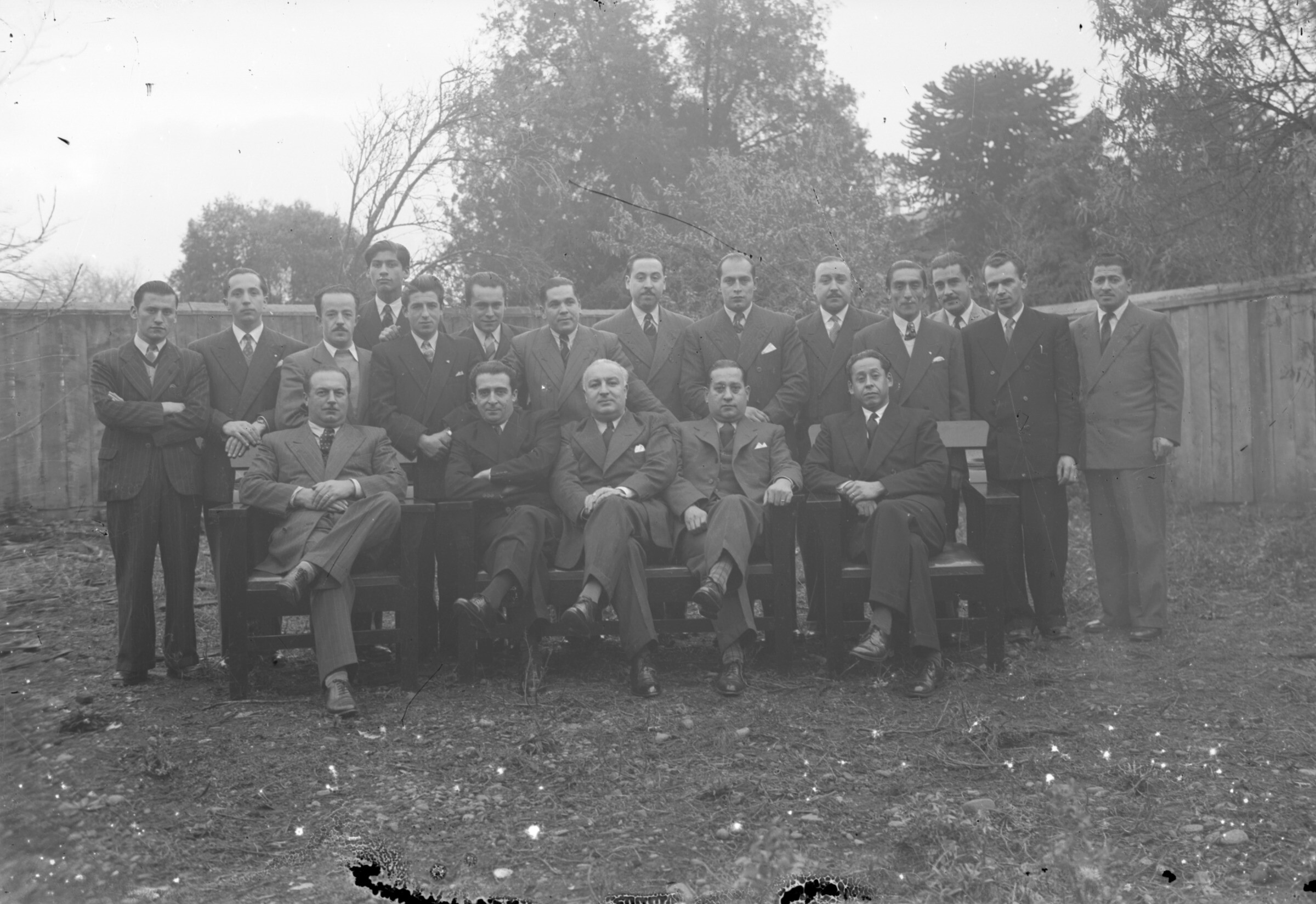 Plano general de un grupo de hombres vestidos formalmente posando de pie o sentados en un patio
