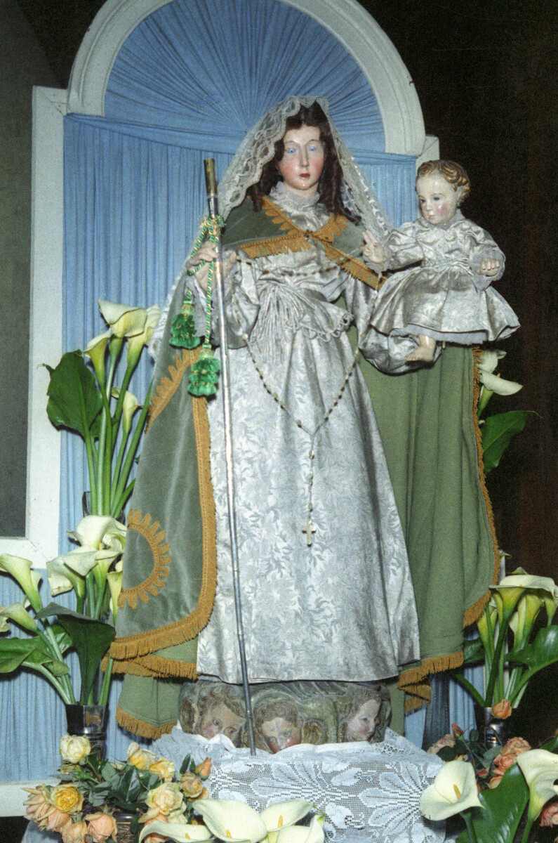 Plano entero de una figura de yeso. Representa la imagen de la virgen María sosteniendo un Bebé en su mano, rodeada de calas sobre el altar de la parroquia San José